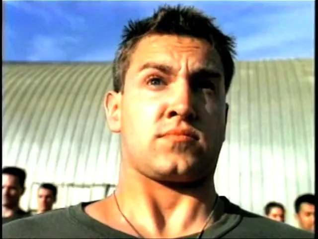 A young man in a green t-shirt looks up with a serious expression, standing outdoors with other men in the background.