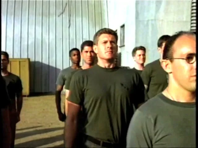 A group of men in military-style t-shirts stand in formation outdoors, with one man in the foreground looking directly ahead.