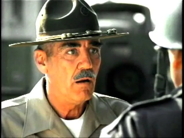 A drill sergeant with a mustache and hat looks surprised while talking to a recruit in an outdoor setting.