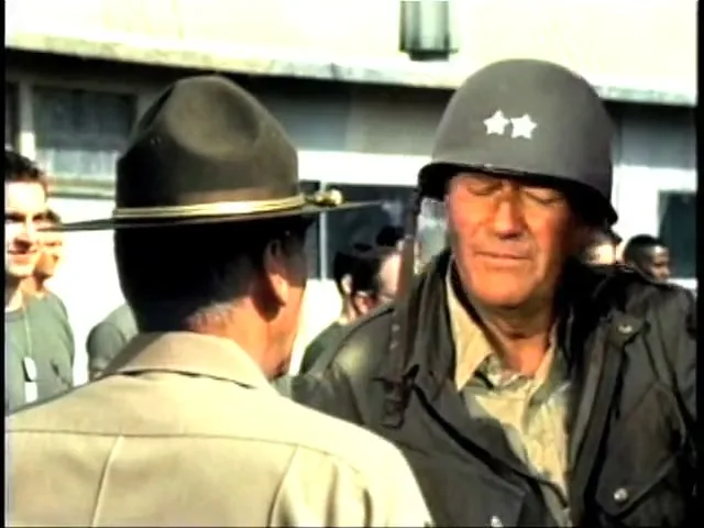 A man in a military helmet with two stars on it talks to another man in a military hat, with other soldiers in the background.