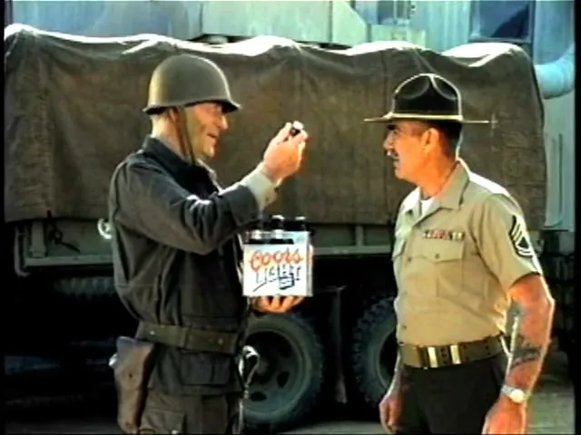 A soldier in a helmet and uniform holds a Coors Light six-pack and a single bottle, showing it to a drill sergeant in a hat and uniform, outdoors.