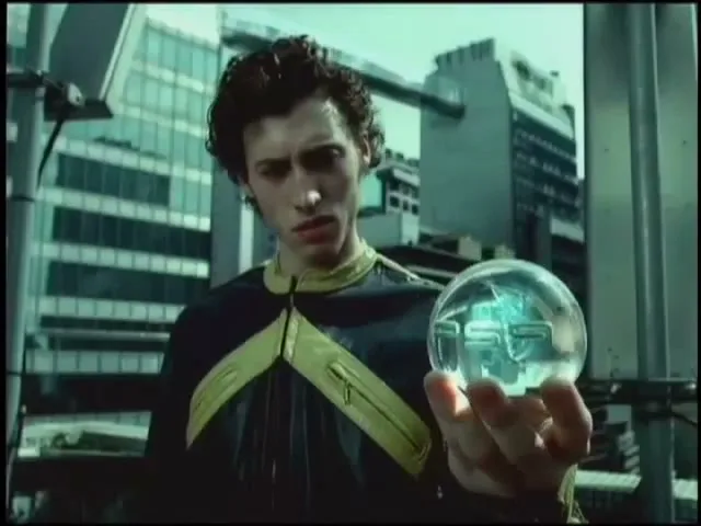 A young man with curly hair in a futuristic jacket holds a glowing orb with "PS9" inside, looking intently at it in a modern city.
