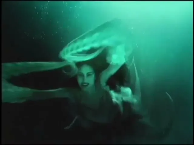 A woman with long hair and dark lipstick swims underwater, looking directly at the camera with a slight smile, as a translucent, jellyfish-like object floats above her.