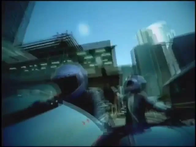 Two people in blue helmets and futuristic suits ride motorcycles through a city street, with buildings reflected in the foreground.