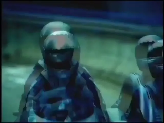 A group of futuristic, armored figures with helmets are seen in a blurry, fast-moving shot, possibly in a virtual city or race.