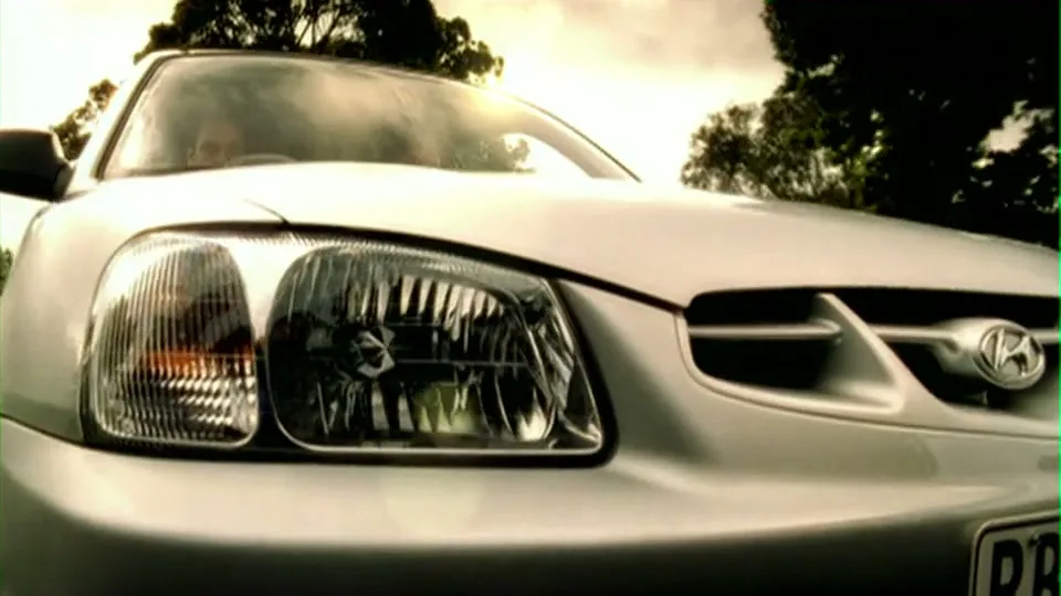 A silver car is parked on a suburban street, with two people visible through the windshield as the camera pulls back.