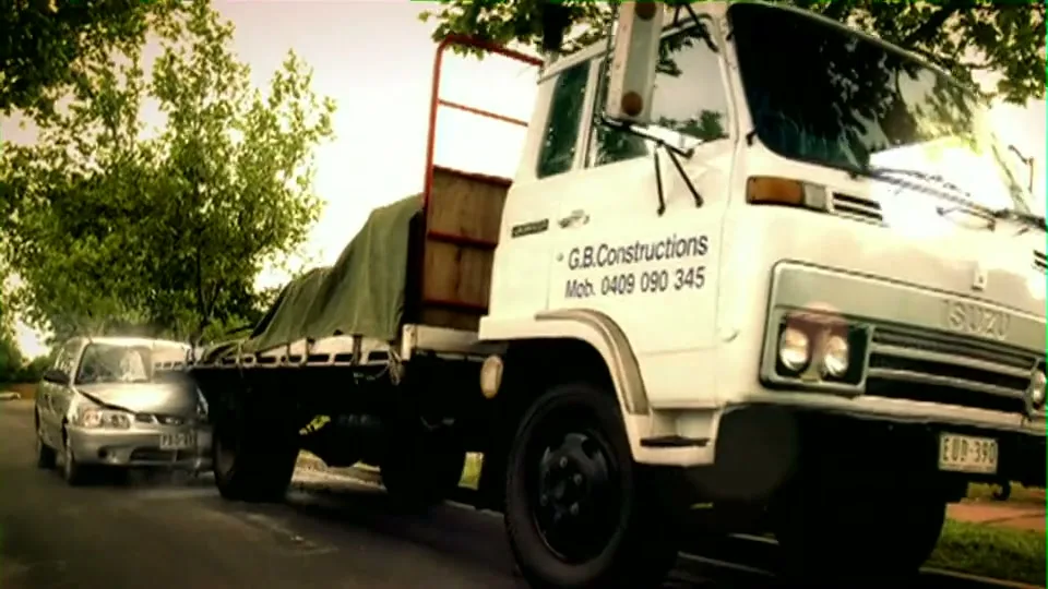 A silver car with a male driver and female passenger crashes into a white flatbed truck on a suburban street during the day.