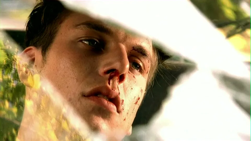 A young man with a bloody nose and face looks up, reflected in a car's broken windshield, with trees visible in the background.