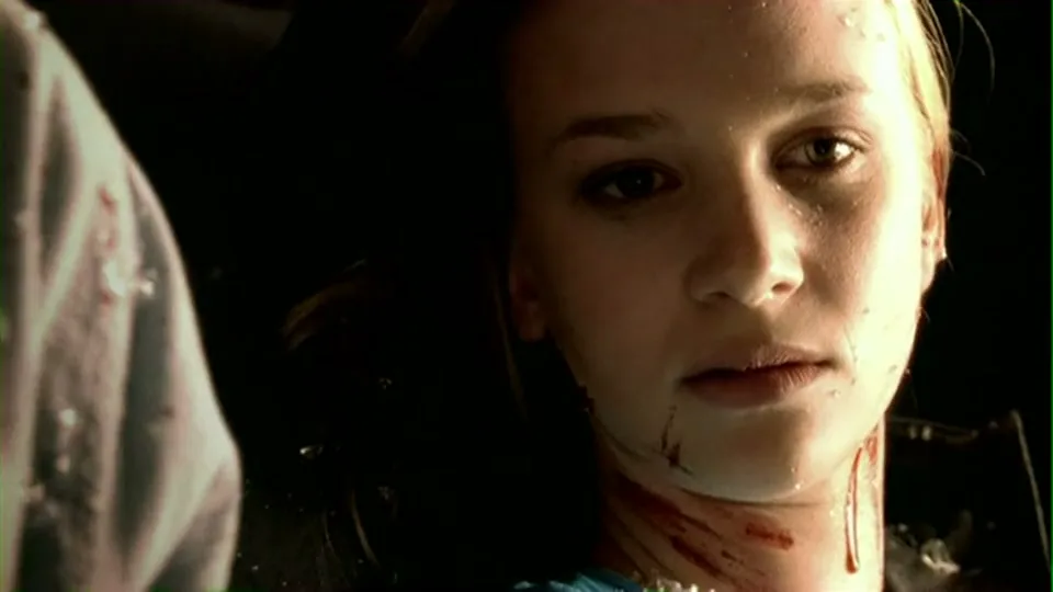 A young woman with blonde hair and blood on her face and neck looks directly at the camera, appearing dazed and injured inside a dark vehicle.