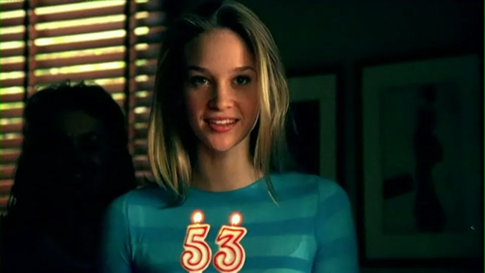 A young woman in a blue striped shirt smiles, holding a birthday cake with "53" candles, in a dimly lit room with blinds.