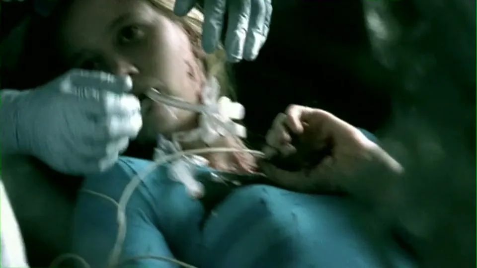 A young woman with blonde hair, wearing a blue hospital gown and intubated, lies on a stretcher as medical personnel attend to her in a dark, chaotic ambulance interior.