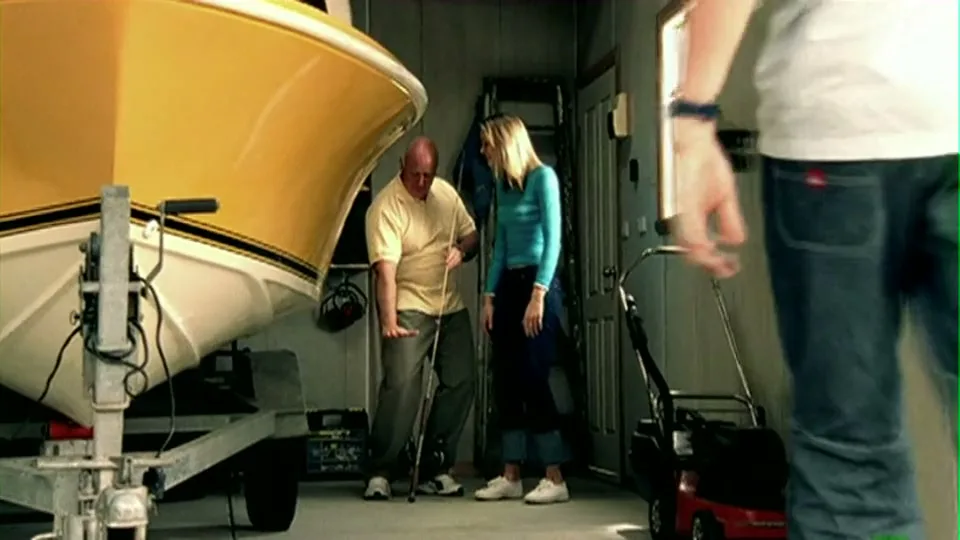 A man with a crutch and a woman talk next to a boat on a trailer in a garage, as a third person walks past them.