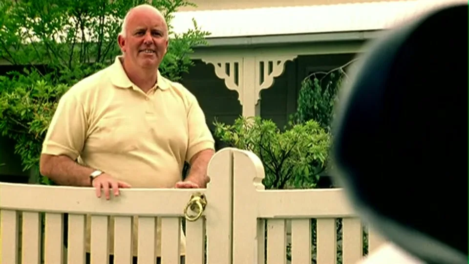 A bald man in a yellow polo shirt leans on a white picket fence, looking at someone off-screen in a suburban setting.