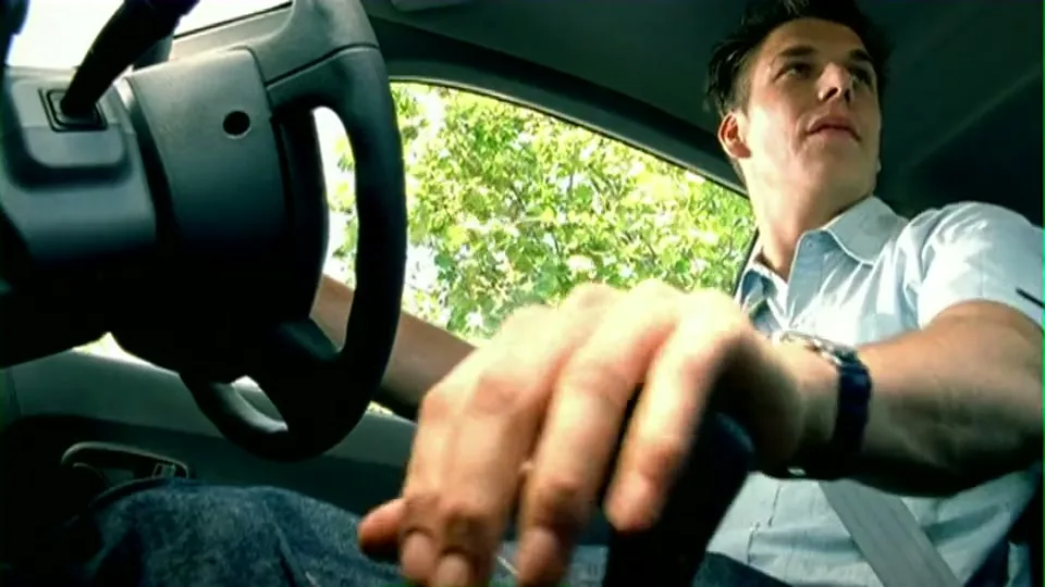 A young man in a light blue shirt and jeans sits in the driver's seat of a car, looking out the window with a concerned expression.