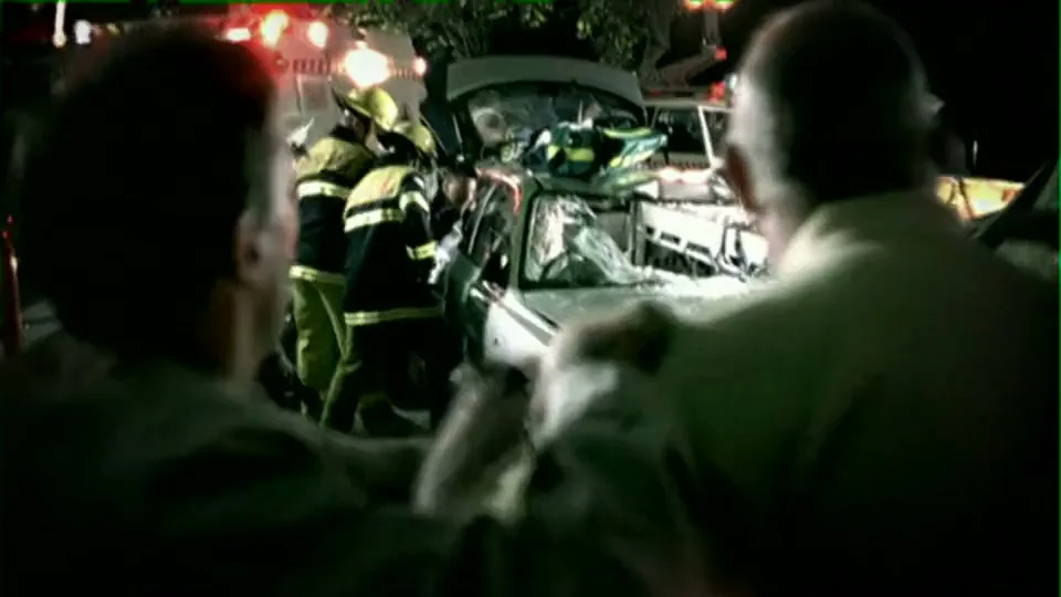 Two men in civilian clothes watch as emergency services, including firefighters and police, attend to a severe car crash at night on a dark road.