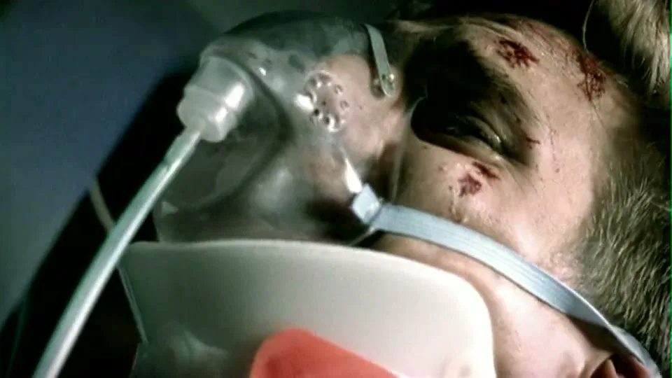 A man with an oxygen mask, neck brace, and blood on his face lies injured, with a red light flashing on him.