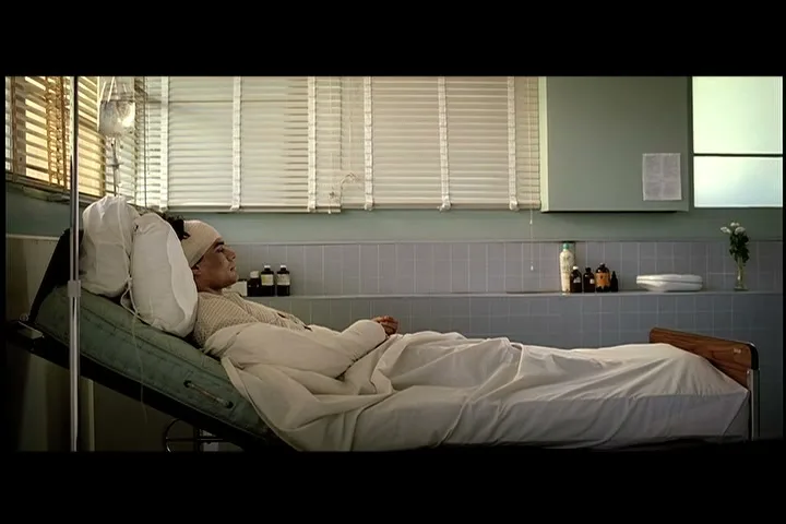A male patient with a bandaged head lies in a hospital bed with an IV drip, looking out a window in a hospital room.