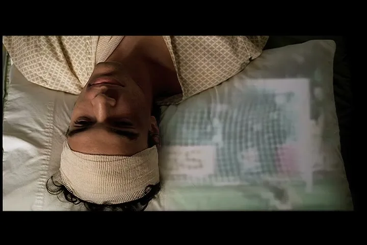 A man with a bandaged head lies in a hospital bed, as images of children's faces are projected onto his pillow.