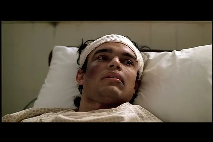 A young man with a bandage on his head and a black eye lies in a hospital bed, looking around with a dazed expression.