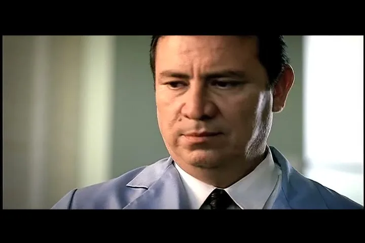 A man with a swollen face, wearing a light blue shirt and tie, looks down and then up, as if in thought, in an interior setting.