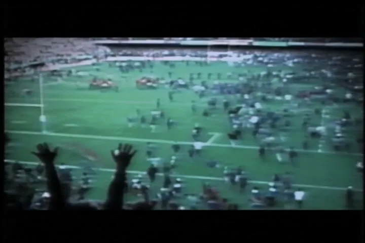 A wide shot from a high angle shows a crowd of people running onto a football field, with some hands raised in the foreground.