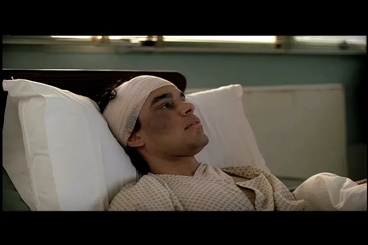A young man with a bandaged head and dirt on his face lies in a hospital bed, looking off-camera with a pensive expression.