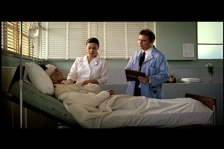 A male patient with bandages on his head lies in a hospital bed, while a female nurse and a male doctor stand beside him in a hospital room.