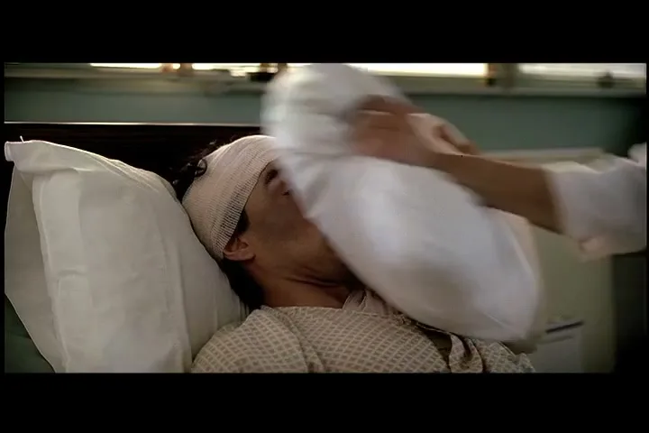 A man with a bandaged head lies in a hospital bed as a pillow is pressed onto his face by an unseen person in a hospital room.