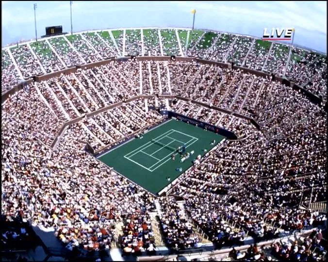 An extreme wide, high-angle shot of a tennis stadium filled with a crowd, with a tennis match in progress on the green court.