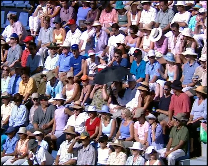A crowd of people, mostly adults, are seated in an outdoor stadium, watching a tennis match on a sunny day.