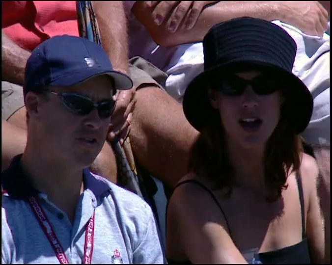 A man in a blue cap and sunglasses sits next to a woman in a black hat and sunglasses, both watching a tennis match in an outdoor stadium.
