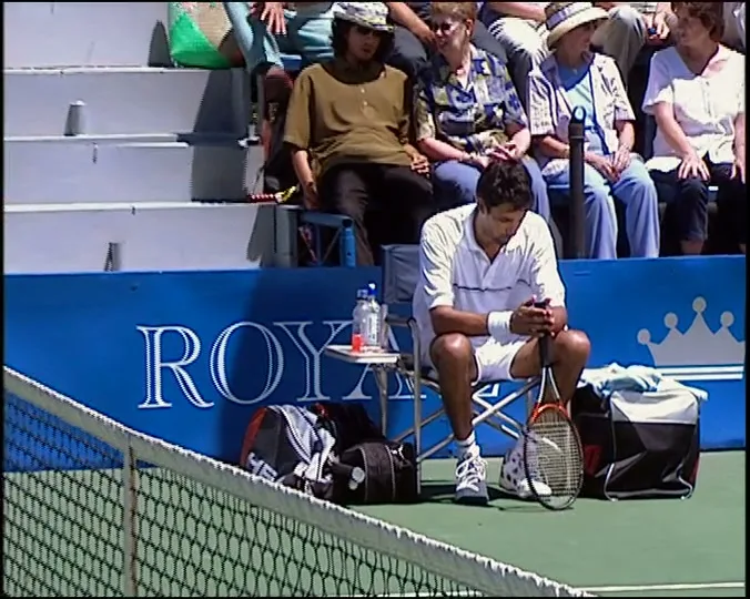 A male tennis player in white sits on a chair on the court, looking around while holding his racket, with spectators in the background.