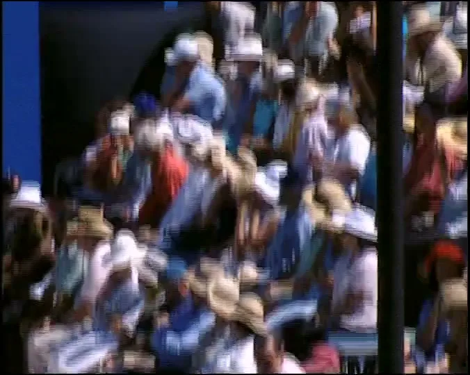 A crowd of people, many wearing hats, are seated in a stadium, watching a tennis match on a sunny day.