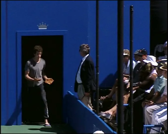 A young man with curly hair, wearing a grey t-shirt and dark pants, walks out of a blue door onto a tennis court, holding a tennis ball and a glove.