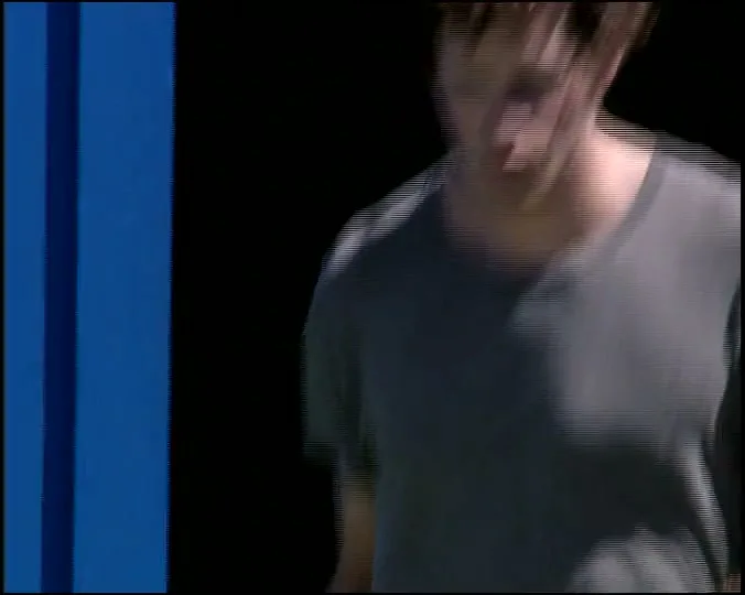 A young man with curly hair in a grey t-shirt walks across a tennis court, looking focused and determined.