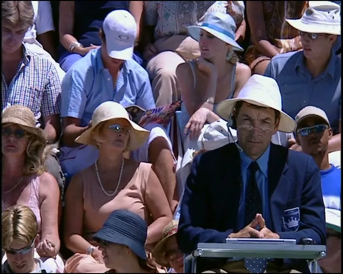 A male tennis umpire in a white hat and blue blazer sits at a table, looking forward, surrounded by a crowd of spectators at a sunny outdoor tennis match.