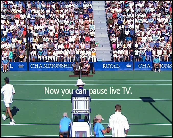 A wide shot of a tennis match with players, a referee, and a large crowd in the stands, with text "Now you can pause live TV." appearing on screen.