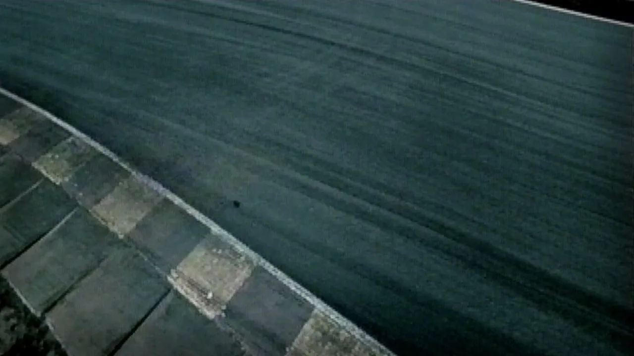 An aerial view shows a dark race track with a checkered curb, as a small, dark object moves quickly across the track.
