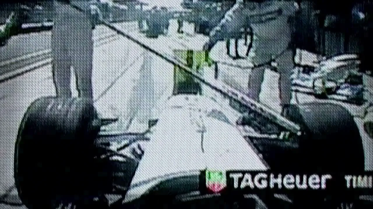 A low-quality, grainy shot from a race car's perspective shows pit crew members working on the car during a pit stop at a sunny racetrack.
