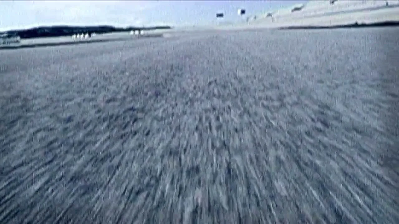A low-angle POV shot from a race car shows the asphalt track blurring past, then the car takes a sharp turn, revealing the colorful curb of the race track.