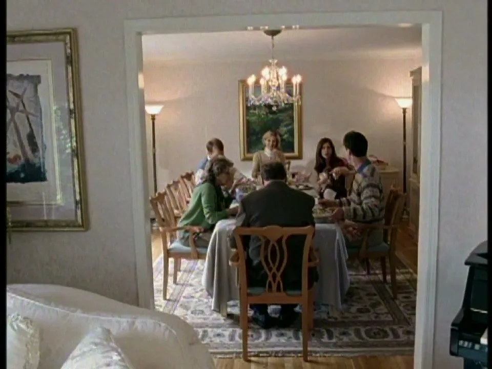 A family of seven people, including children and adults, are seated around a dining table in a brightly lit dining room, eating a meal.