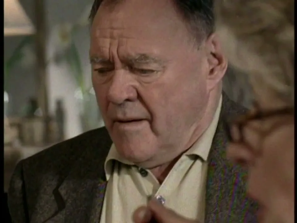 A man in his 60s, wearing a brown jacket and light shirt, looks down as a woman's hand gestures in front of him in an interior setting.