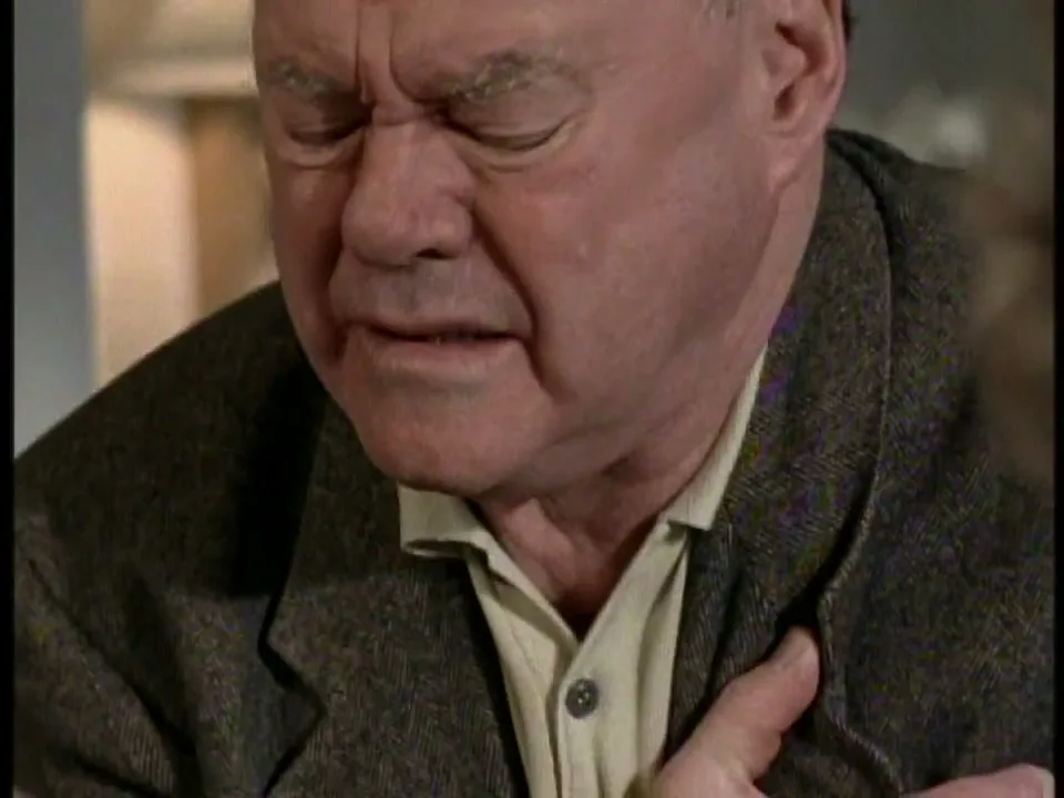 A distressed older man in a brown jacket clutches his chest, eyes closed in pain, in an interior setting.