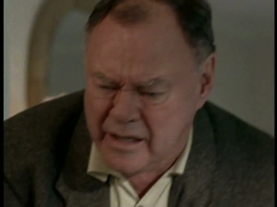 A man in his 60s, wearing a suit jacket and collared shirt, grimaces in pain or discomfort in an interior setting.