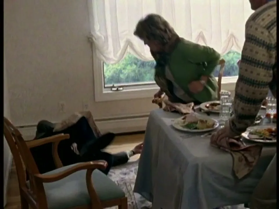 A woman in a green cardigan and scarf leans over a dining table, looking down at a man on the floor, while another man stands at the table.