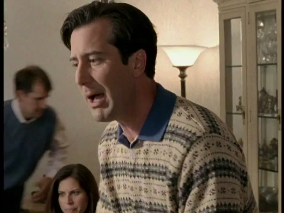 A man in a patterned sweater and blue polo shirt talks to someone off-screen, while a woman and another man are blurred in the background at a family gathering.