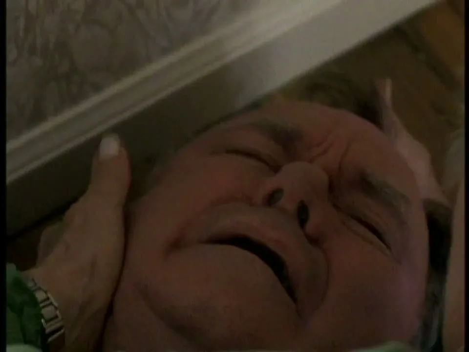 A man with gray hair and a green blanket is lying on the floor, grimacing in pain as a woman's hands hold his head.