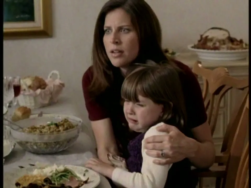 A woman in a maroon shirt holds a crying child at a dining table, looking off-screen with a worried expression during a Thanksgiving meal.