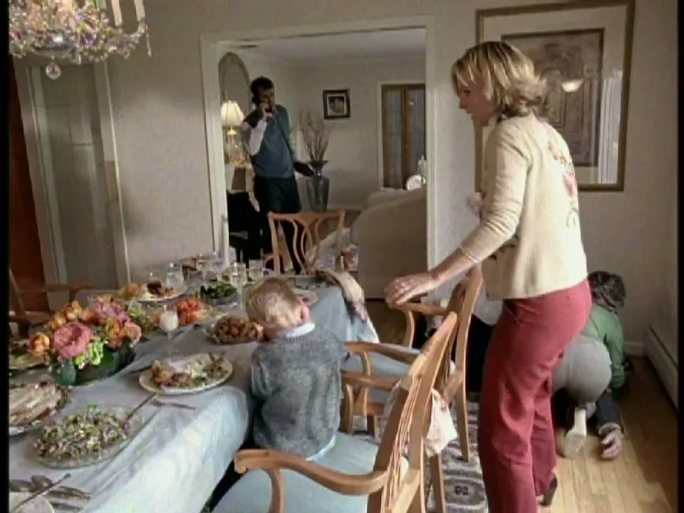 A woman in a beige cardigan and red pants stands at a dining table, looking concerned, while a man talks on the phone in the background and children play on the floor.