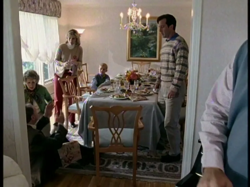 A family gathers around a dining table, looking down at a man on the floor, while another man talks on the phone in the foreground.